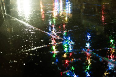 Mavi, yeşil ve kırmızı Noel ışıkları yağmurlu bir gecede kaldırıma yansıyor.
