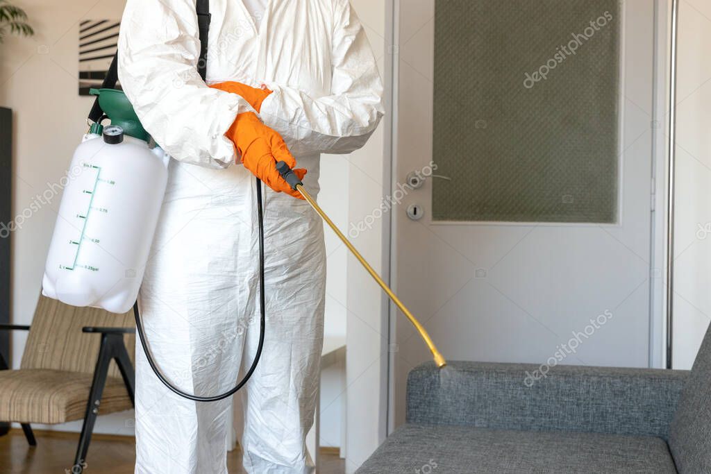 Person in protective suit with decontamination sprayer bottle disinfecting household and furniture