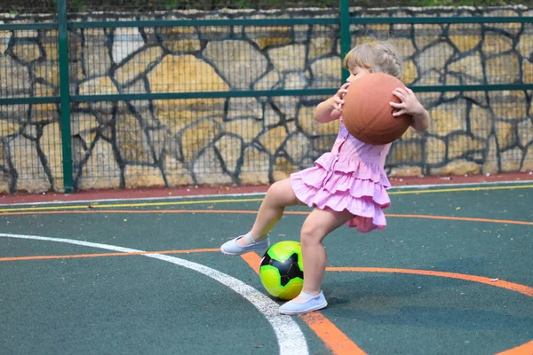 小さな女の子がスポーツ場にボールで落ちる。大きなバスケットボールをしている子供 — ストック写真