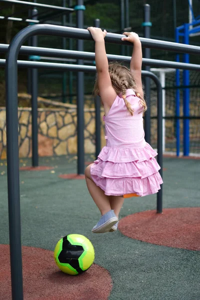 Niña niña cuelga en barra horizontal en campo de deportes al aire libre. — Foto de Stock