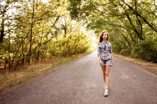 Krásná dívka chůze na prázdné silnici mezi zelenými stromy — Stock fotografie