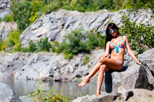 Beautiful  girl in a colorful bikini sits on a rock near a mount — Stock Photo, Image