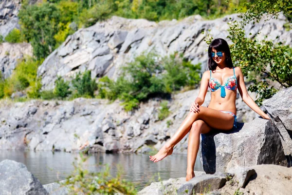Tanned brunette girl in a colorful bikini sits on a rock near a — Stock Photo, Image