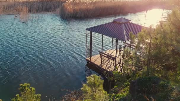 日没時に川の上にガゼボと小さな木製の桟橋 葦と火の間の池の上に空のアーバー ない人 リラックスした美しい水のシーンのための場所 — ストック動画