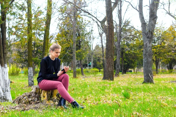 Gyönyörű lány ül a csonkot, a parkban, smartphone — Stock Fotó