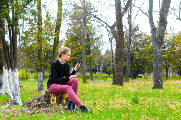 あなたのスマート フォンで公園の切り株の上に座って美しい少女 — ストック写真