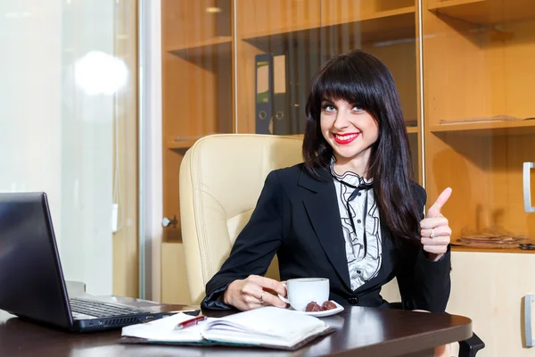 Hermosa mujer sonriente en la oficina bebiendo café y muestra thum — Foto de Stock