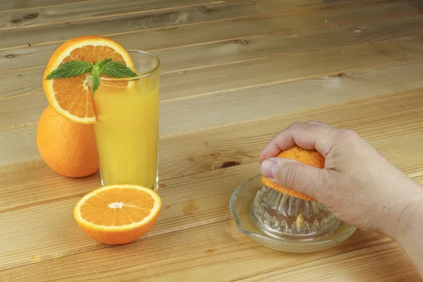 A hand squeezing juice from an orange on a manual glass squeezer. Set on a wooden planked table. Domestic production of orange juice. — Stock Photo, Image