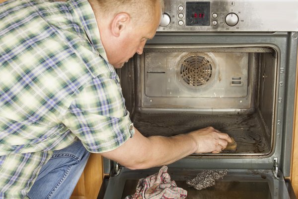 Мужчина преклоняет колени на полу на кухне и чистит духовку. Уборка дома. Мужчина помогает своей жене со служанкой
.