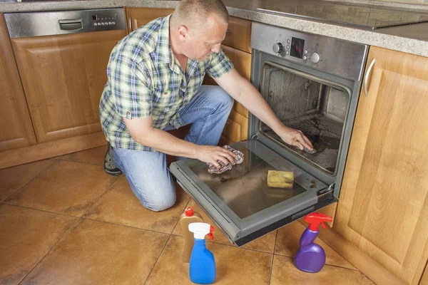 O homem ajoelha-se no chão da cozinha e limpa o forno. Trabalho de limpeza em casa. Homem ajudando sua esposa com serviço de empregada doméstica . — Fotografia de Stock