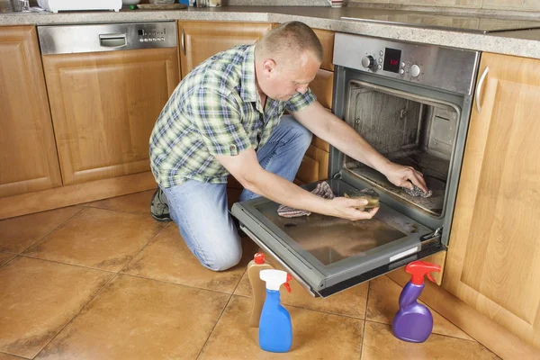 El hombre se arrodilla en el suelo de la cocina y limpia el horno. Trabajos de limpieza en el hogar. Hombre ayudando a su esposa con el servicio de limpieza . —  Fotos de Stock