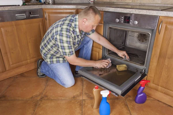 Man kneels on the floor in the kitchen and cleans the oven. Cleaning work in the home. Man helping his wife with maid service. — Stock Photo, Image