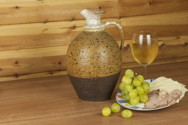 Old clay jug and a glass of wine on a wooden table. White wine and snacks. Ham, cheese and grapes to eat. Relax with wine and good food. Place for your text. Wooden blurred background.