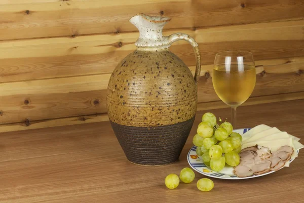 Old clay jug and a glass of wine on a wooden table. White wine and snacks. Ham, cheese and grapes to eat. Relax with wine and good food. Place for your text. Wooden blurred background. — Stock Fotó