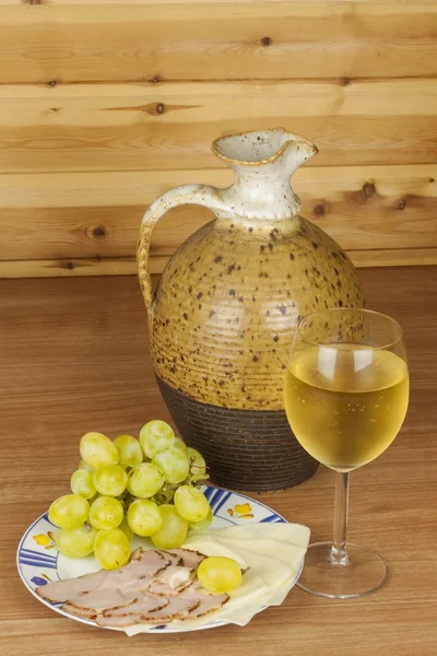 Old clay jug and a glass of wine on a wooden table. White wine and snacks. Ham, cheese and grapes to eat. Relax with wine and good food. Place for your text. Wooden blurred background. — Stock Fotó