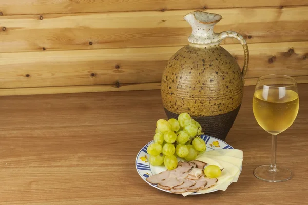 Old clay jug and a glass of wine on a wooden table. White wine and snacks. Ham, cheese and grapes to eat. Relax with wine and good food. Place for your text. Wooden blurred background. — Stock Fotó