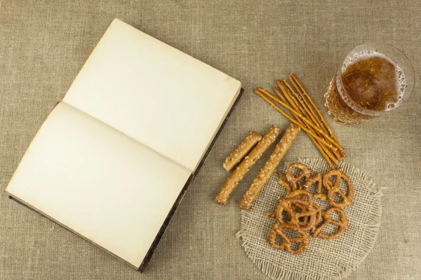 Cerveza en un vaso y un libro sobre la mesa. Paja de trigo crujiente con sal. Montón de palitos de pretzel. Entretenimiento para ver TV . —  Fotos de Stock