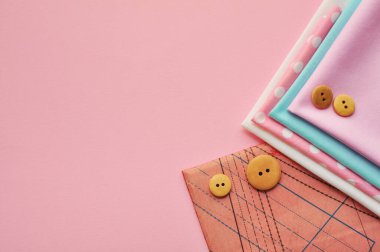 Farklı renklerde pamuk kumaş, desenler, renkli kurdeleler ve pembe arka planda ahşap düğmeler. Metin için boşluk, üst görünüm 