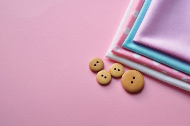 Farklı renklerde pamuk kumaş, desenler, renkli kurdeleler ve pembe arka planda ahşap düğmeler. Metin için boşluk, üst görünüm 