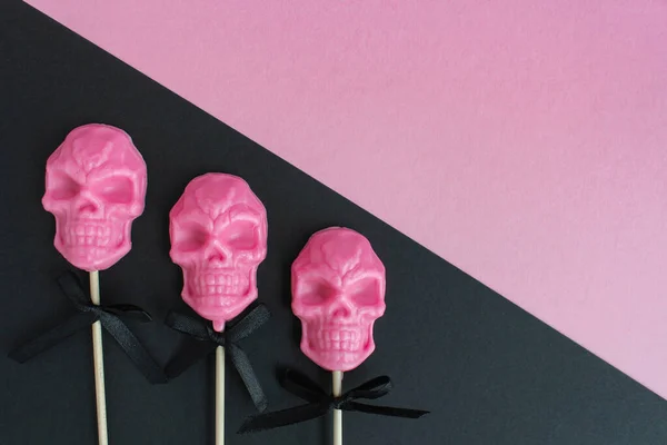 three pink lollipops in the shape of a skull on a stick on a pink and black background. the concept of Halloween.