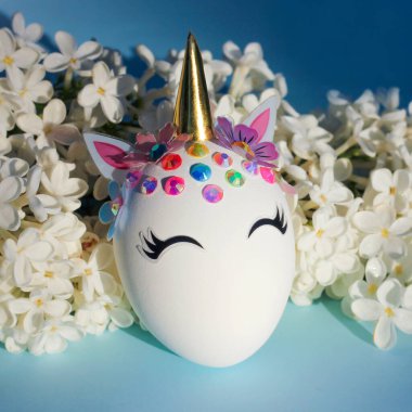 Tek boynuzlu at şeklinde yaratıcı bir Paskalya yumurtası. Mavi arka planda beyaz narin çiçekler var.