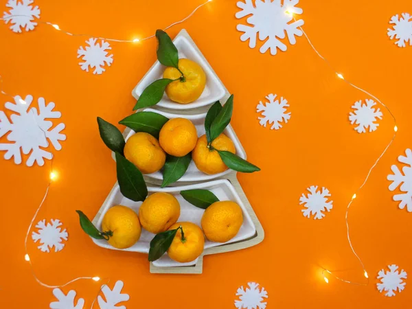Turuncu zemin üzerinde beyaz dekoratif kar taneleriyle Noel ağacı şeklinde mandalinalar. Noel yemekleri arka planı, en iyi manzara. Eğlenceli bir Noel ağacı.