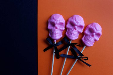Turuncu ve siyah arkaplandaki çubuk üzerinde kafatası şeklinde üç pembe lolipop. Cadılar Bayramı konsepti.