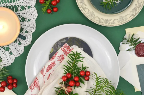 Kırmızı böğürtlen ve yemyeşil dallarla süslenmiş Noel sofra takımı. Retro peçeteler, dekoratif tabaklar, kaplamalı zarflar ve yanan mum..