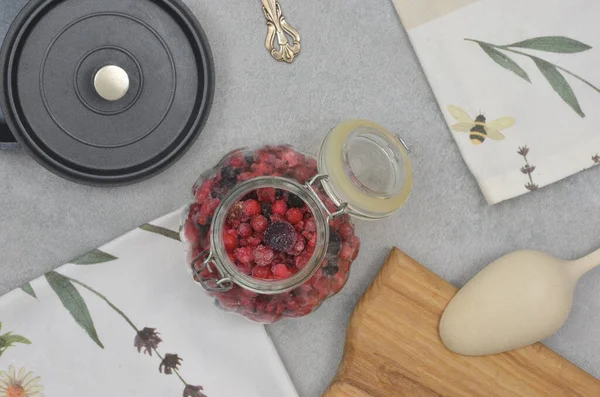 Çilekli süsleme kavanozu, mutfak havlusu, kaşık, siyah tencere kapağı ve mermer masa üzerinde kesim tahtası..