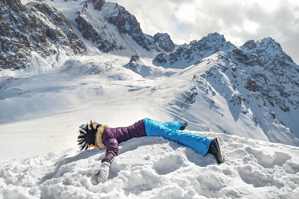 Acrophobia woman in funny mohawk hat lie on top of a snowy rock cliff edge. Fighting the fear of heights concept.