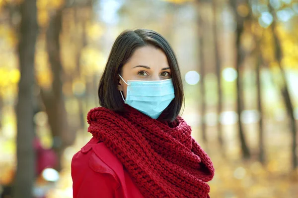Beautiful young woman in medical protective face mask walking outdoors in autumn park. Covid-19 quarantine concept
