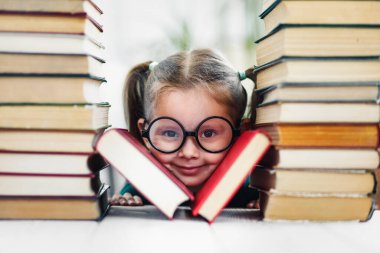 Anaokulu çağında gamzeli ve kocaman komik gözlüklü bir kız kitap yığınının arasında gülümsüyor.