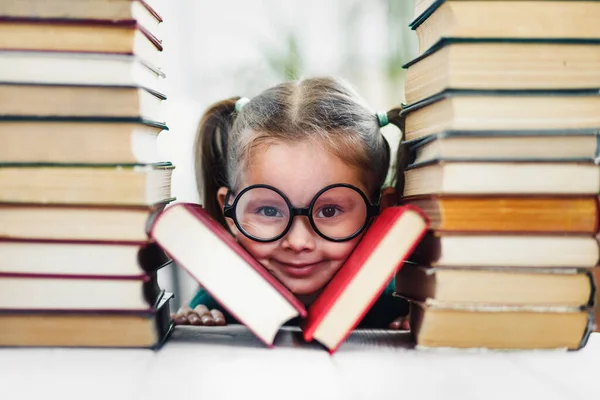 Preschool age smiling girl with dimples in big funny glasses among a pile of books — Stock Photo, Image