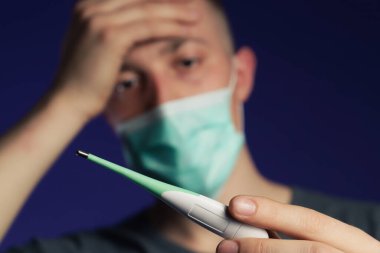 Kafasında avuç içi termometreyle tıbbi gazlı bez maskeli beyaz bir adam, ateş kavramı.