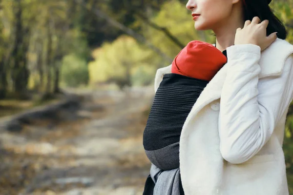 Hermosa madre joven con su bebé en honda al aire libre. La mujer lleva a su hijo y camina en otoño por la naturaleza. Concepto de bebé —  Fotos de Stock