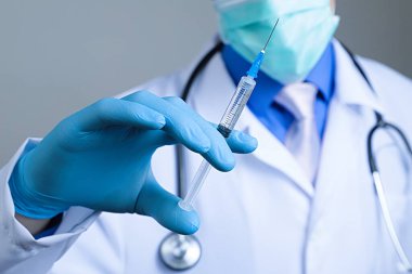 Plastik mavi eldivenli tıbbi şırıngaya yakından bak. El giydirilmiş, göğsü beyaz tıbbi bornozlu, steteskoplu, bulanık bir arka planı var.