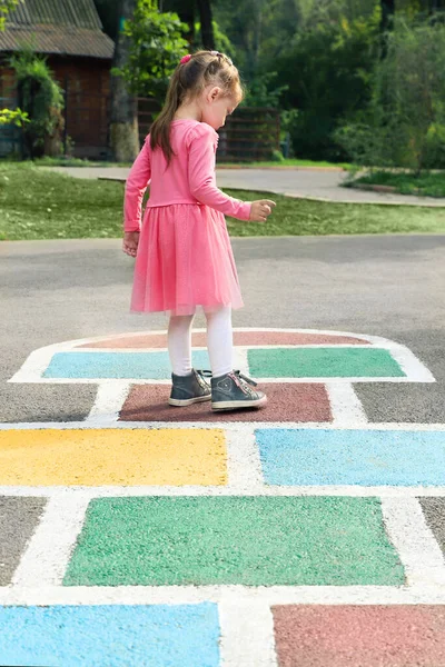 Dziewczynka w różowej sukience bawiąca się w hopscotch na placu zabaw na świeżym powietrzu, zajęcia na świeżym powietrzu dla dzieci. — Zdjęcie stockowe