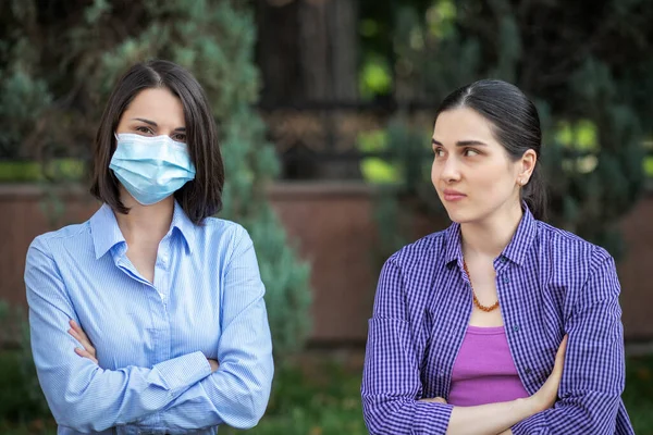 Дві молоді жінки сидять на парковій лавці. Одна жінка в масці, одна без маски. Різні ставлення до пандемії . — стокове фото