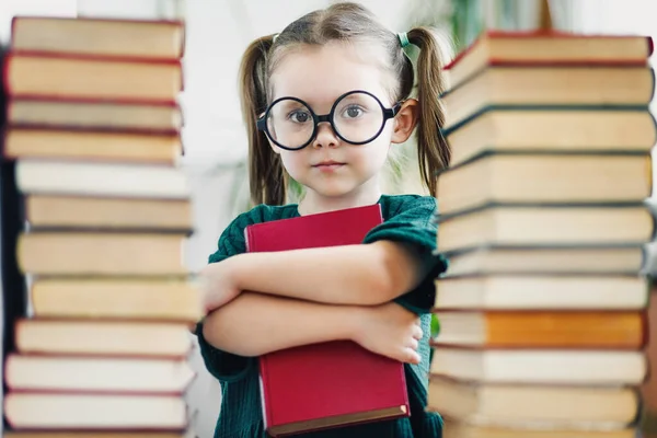 Preschool age girl in big round glasses holding red book among book piles — Stock Photo, Image