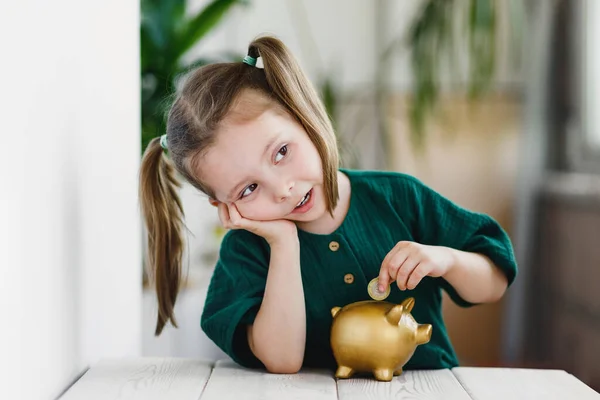 Krásná roztomilá dívka dávat peníze do zlaté prasátko banky u stolu uvnitř. Primární finanční vzdělávání — Stock fotografie