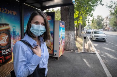 Genç bir kadın, covid-19 salgını sırasında antivirüs maskeli bir durakta durup otobüs bekliyor.