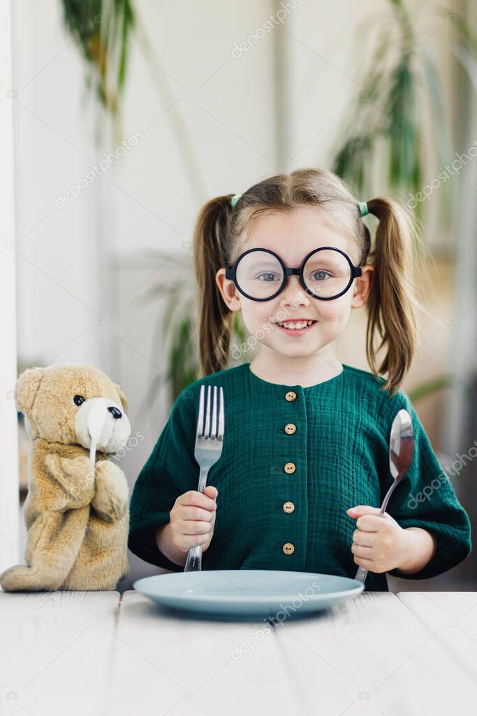 Little beautiful girl in green muslin dress waiting breakfast with bear toy.