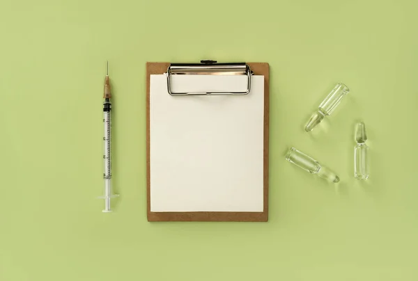 Blank medical clipboard, syringe and ampoules with transparent liquid on green background. Natural medicine background. Coronavirus vaccine concept.