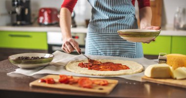Önlüklü bir kadın şef çiğ pizzaya domates sosu koyuyor. Evde, mutfakta lezzetli pizzalar pişirmek..