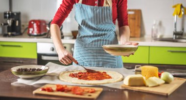Önlüklü bir kadın şef çiğ pizzaya domates sosu koyuyor. Evde, mutfakta lezzetli pizzalar pişirmek..