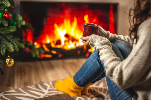 Woman in a sweater and warm socks is sitting in front of the fireplace, which is depicted on the TV screen and drinking a hot beverage from a cup. Concept of creating a cozy winter atmosphere at home