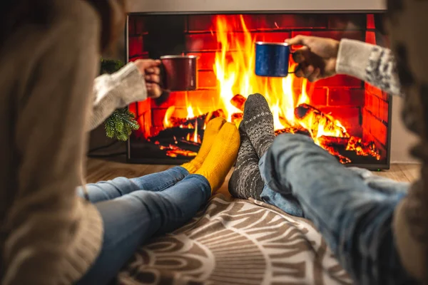 Man and woman are sitting next to the fireplace, which is depicted on the TV screen and drinking a hot beverage. Concept of creating a cozy winter atmosphere at home