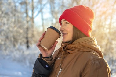 Genç bir kadın soğuk bir kış gününde sıcak kahve içiyor. Açık havada, ormanda güneşli bir günde.