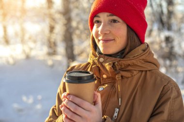 Genç bir kadın soğuk bir kış gününde sıcak kahve içiyor. Açık havada, ormanda güneşli bir günde.