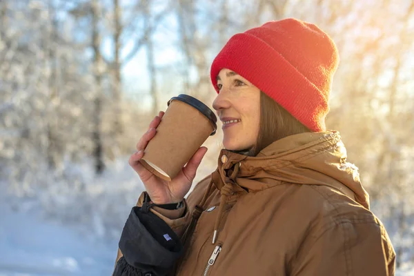 Genç bir kadın soğuk bir kış gününde sıcak kahve içiyor. Açık havada, ormanda güneşli bir günde.